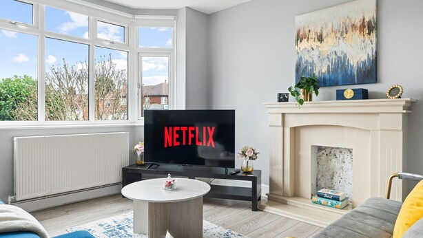 Ein Wohnzimmer mit Kamin, einem Fernseher, der 'NETFLIX' anzeigt, und Blick ins Freie durch große Fenster.