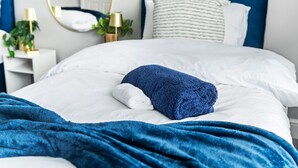 Ein ordentlich bezogenes Bett mit weißer und blauer Bettwäsche, einer blauen Tagesdecke sowie ein Nachttisch mit Lampe und Pflanze.