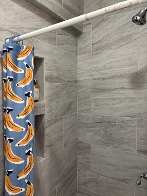 Zona de ducha con azulejos de mármol gris, un cabezal de ducha, una barra para cortina blanca y una botella de gel de ducha.