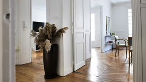A hallway with a wooden floor, white doors, a black vase with dried plants, and a dining area with chairs and a table. - Beaut. Hausmannian flat in Oberkampf near Marais (Paris)