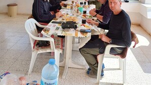 Um grupo de pessoas jantando à mesa com vista para árvores e pássaros do lado de fora.