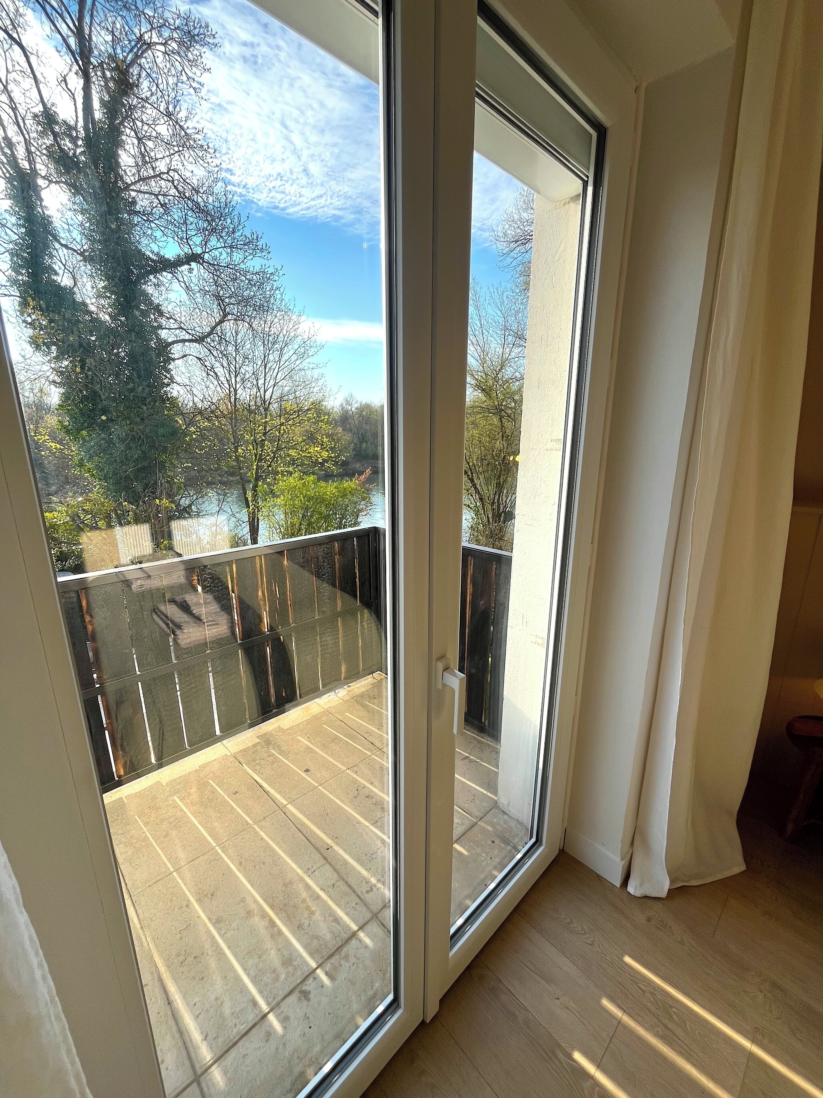 Una habitación con un amplio ventanal corredera que da a un balcón exterior con vistas a árboles y un cuerpo de agua.