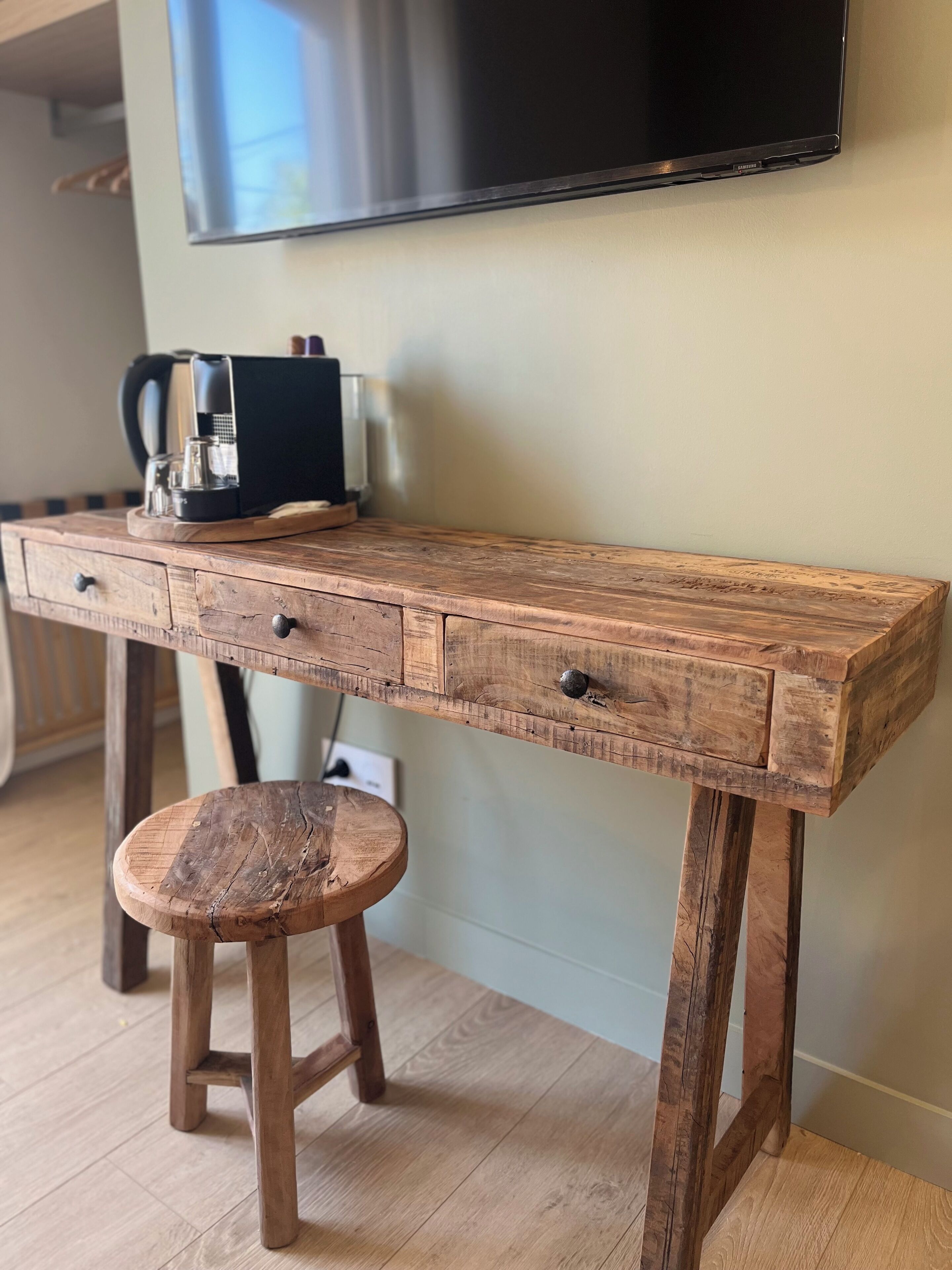 A wooden desk with drawers and a stool, a coffee maker, and a television mounted on the wall.