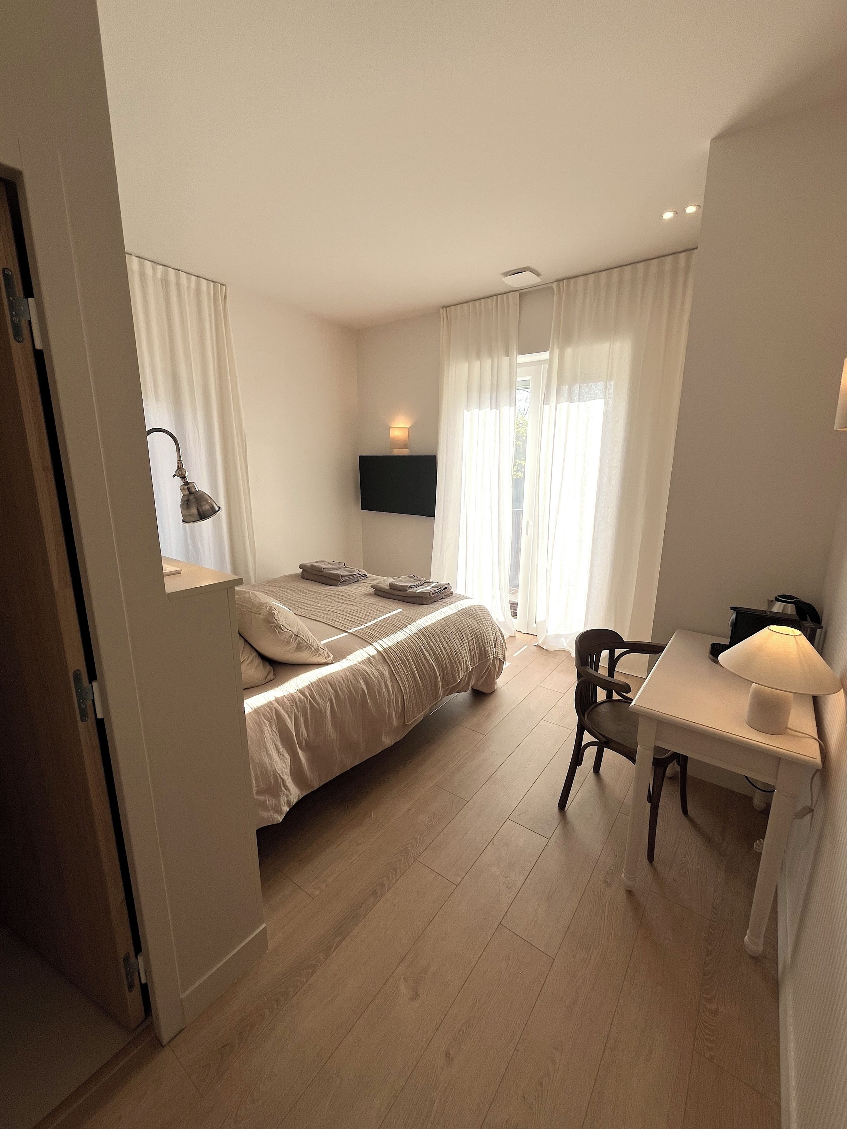 A modern hotel room with a bed, a desk, a chair, a flat-screen TV, and a window with curtains.