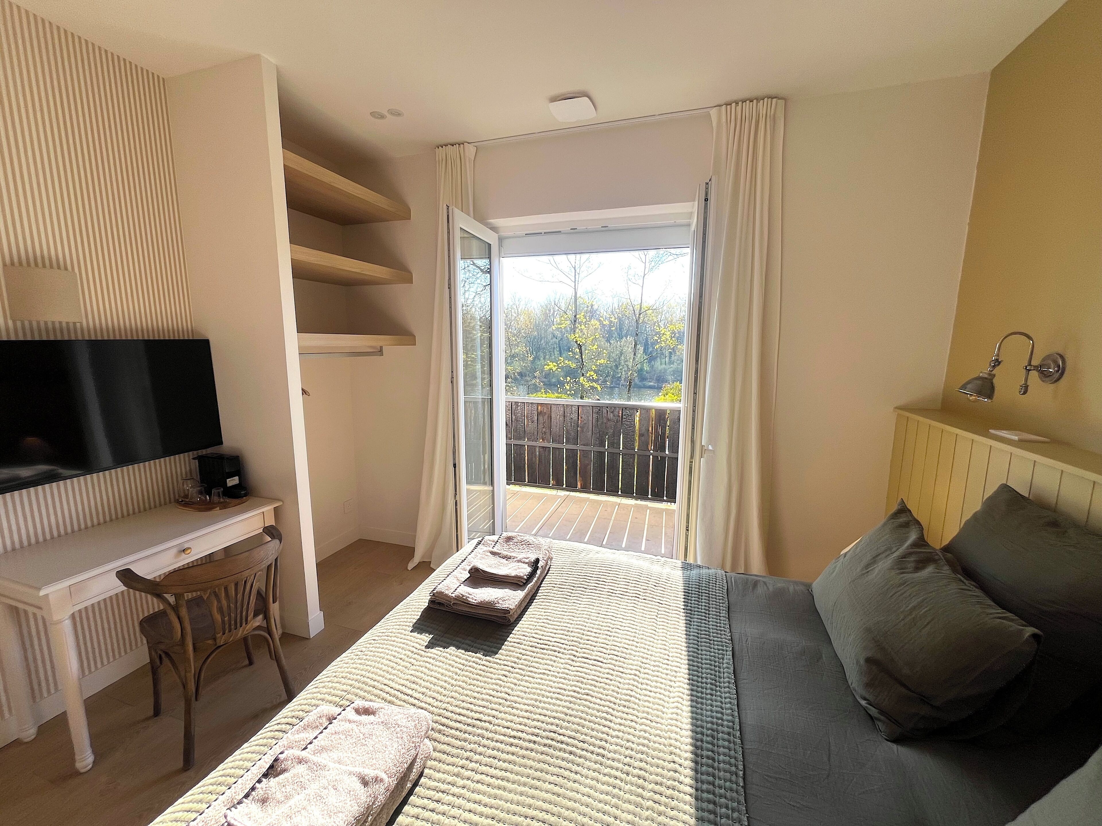 Un dormitorio con cama, escritorio, televisor y balcón con vista a árboles.
