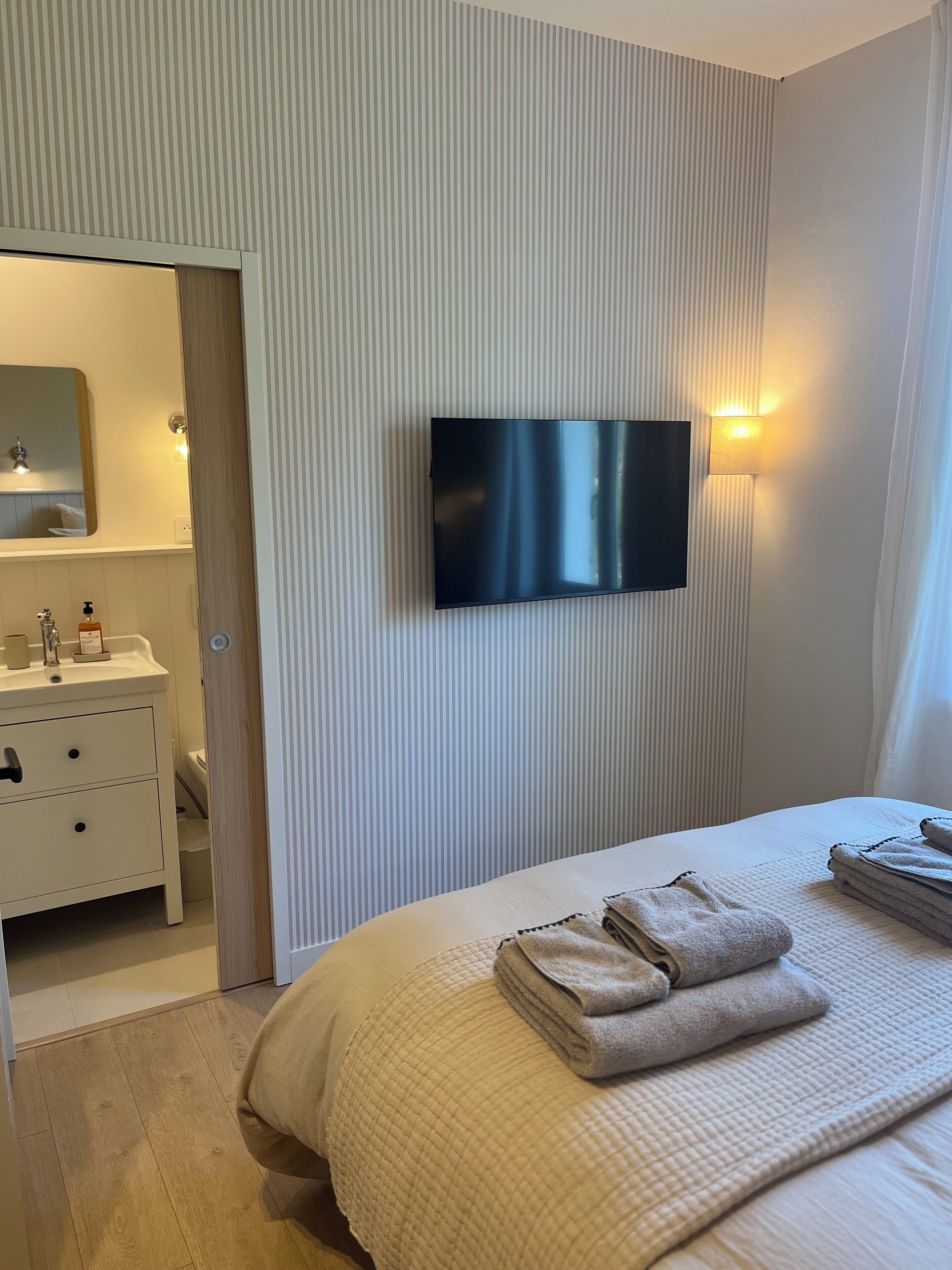 Una habitación de hotel con una cama, un televisor montado en la pared, un baño con lavabo y espejo, y una puerta que da a otra habitación.