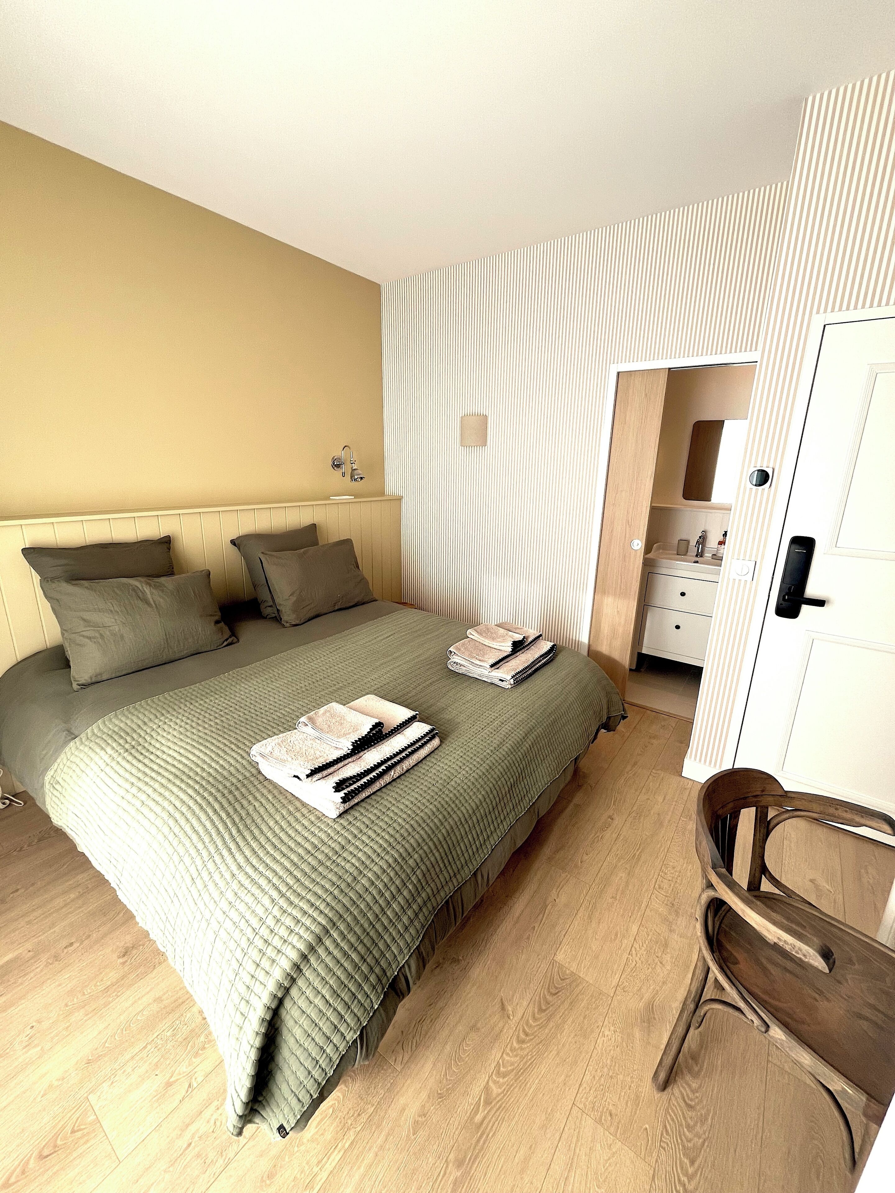 Ein Schlafzimmer mit Bett, Kissen, einem Sessel, einem Nachttisch und einer Tür zu einem Badezimmer.