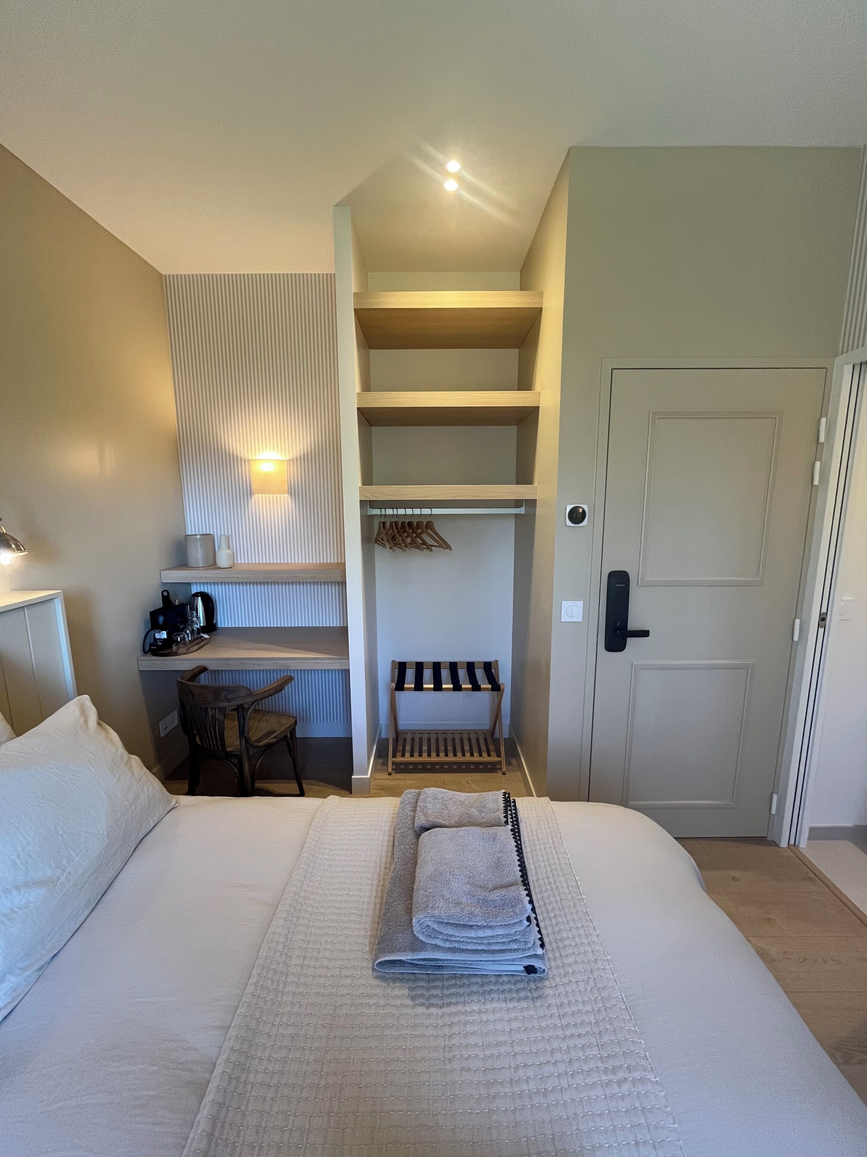 A hotel room with a bed, a desk with a lamp, a chair, a coat rack, and a door.