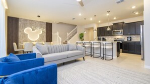 Een moderne woonkamer met een grijze bank, blauwe stoelen, een keuken met donkere kasten en een trap.