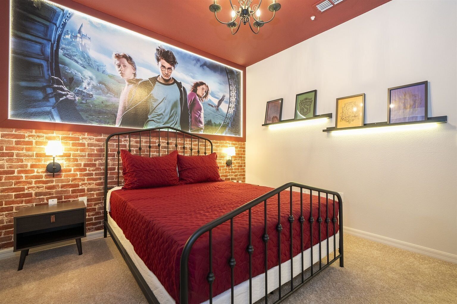 A bedroom with a brick-patterned wall, a metal bed frame, a red bedspread, a nightstand with a lamp, and a large poster of characters on the wall.