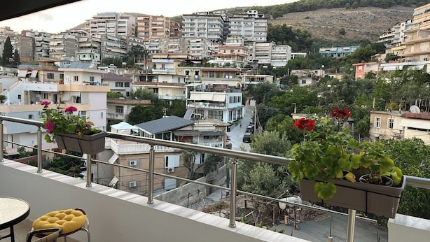 A balcony with potted plants and a table, overlooking a densely built urban area with various buildings and a hillside in the background. - Albania Dream Saranda Apartments (Sarandë)