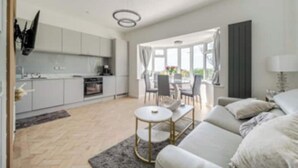 Ein modernes Wohnzimmer mit Sofa und Couchtisch, eine Küche mit grauen Schränken sowie ein Essbereich mit Tisch und Stühlen.