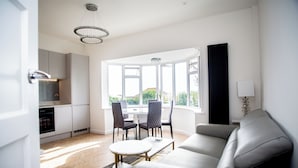 Ein modernes Wohnzimmer mit einer Couch, einem Esstisch mit Stühlen, einer Küchenzeile und großen Fenstern, die für natürliches Licht sorgen.