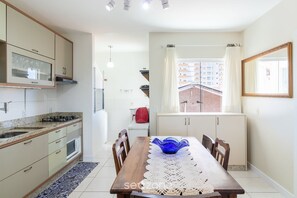 A modern kitchen with a dining area, including a table with a blue bowl, wooden chairs, and a view of a building through a window. - Apt a 600m da praia prox. do Beto Carrero GOT0201 (Penha)