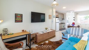 Un salon moderne avec un canapé, une table basse en bois, un téléviseur fixé au mur et une cuisine en arrière-plan.