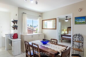 A dining area with a wooden table, chairs, and a blue bowl. There is a white cabinet with a red bag on top. A window with curtains is visible, and a bed with a lamp is seen in the background. - Apt a 600m da praia prox. do Beto Carrero GOT0201 (Penha)