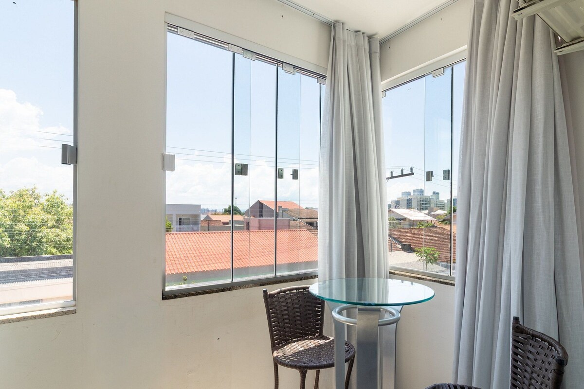 Un balcon avec une table en verre et deux fauteuils en osier, donnant sur un paysage urbain avec des bâtiments aux toits rouges.