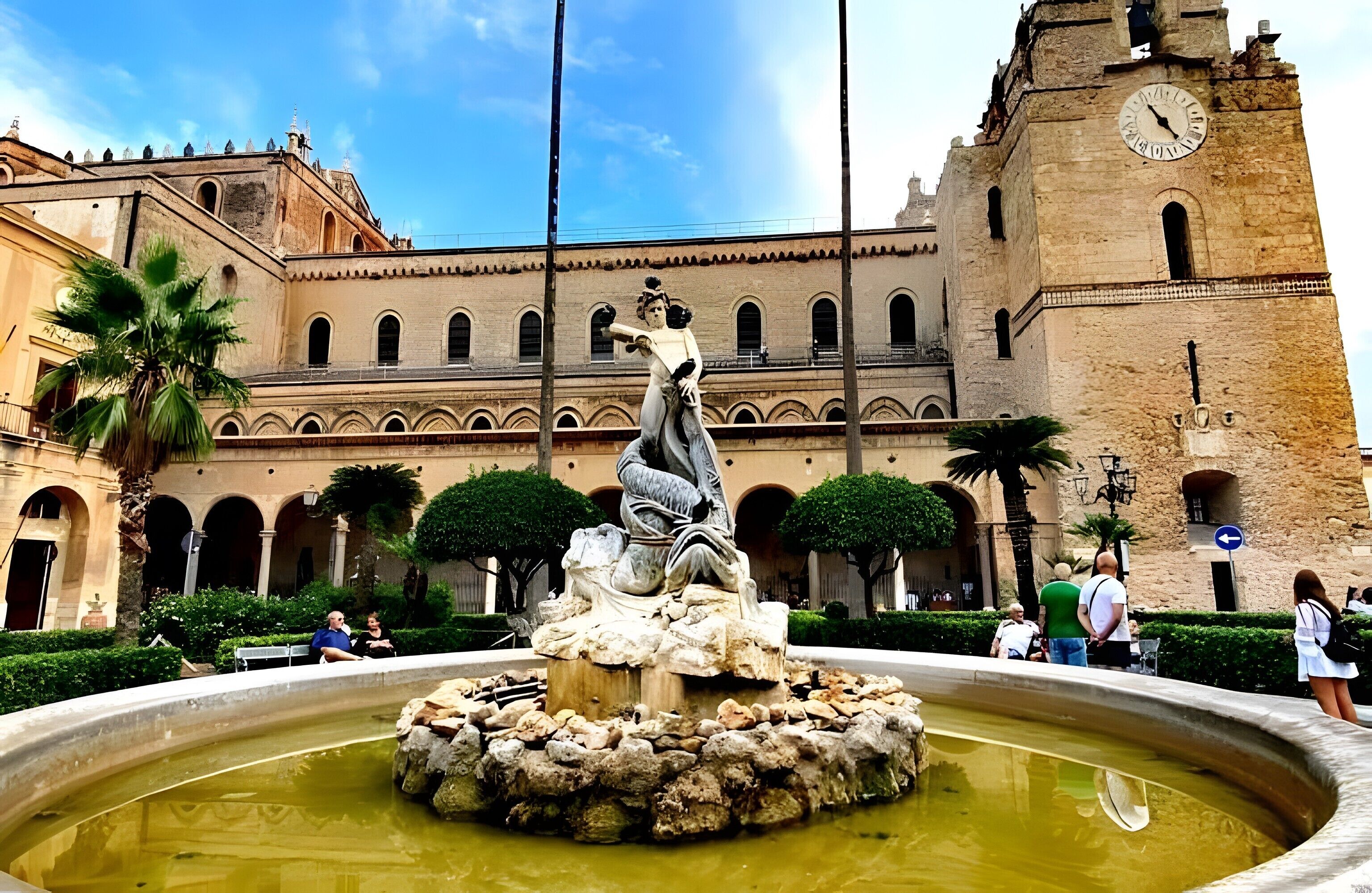 A historic building with a clock tower, a fountain with a statue, and a courtyard with palm trees.