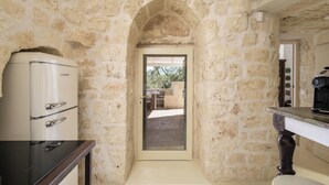 A stone-walled kitchen with a refrigerator, a coffee machine, and a view through an archway to an outdoor area. - Trullo Cecapesce by Perle di Puglia (Carovigno)