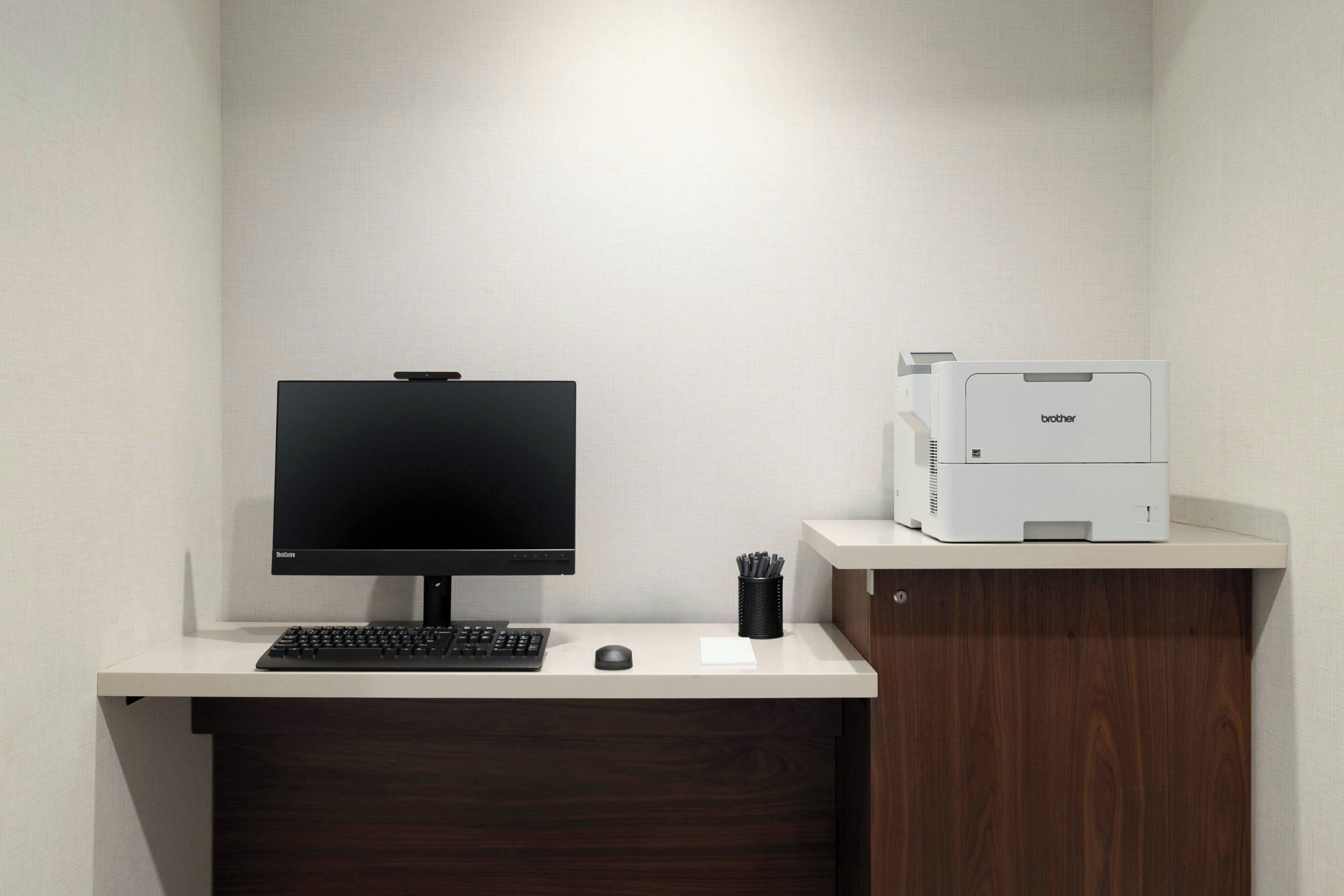 A modern office desk with a computer monitor, keyboard, mouse, and a Brother printer on a white desk against a plain wall.