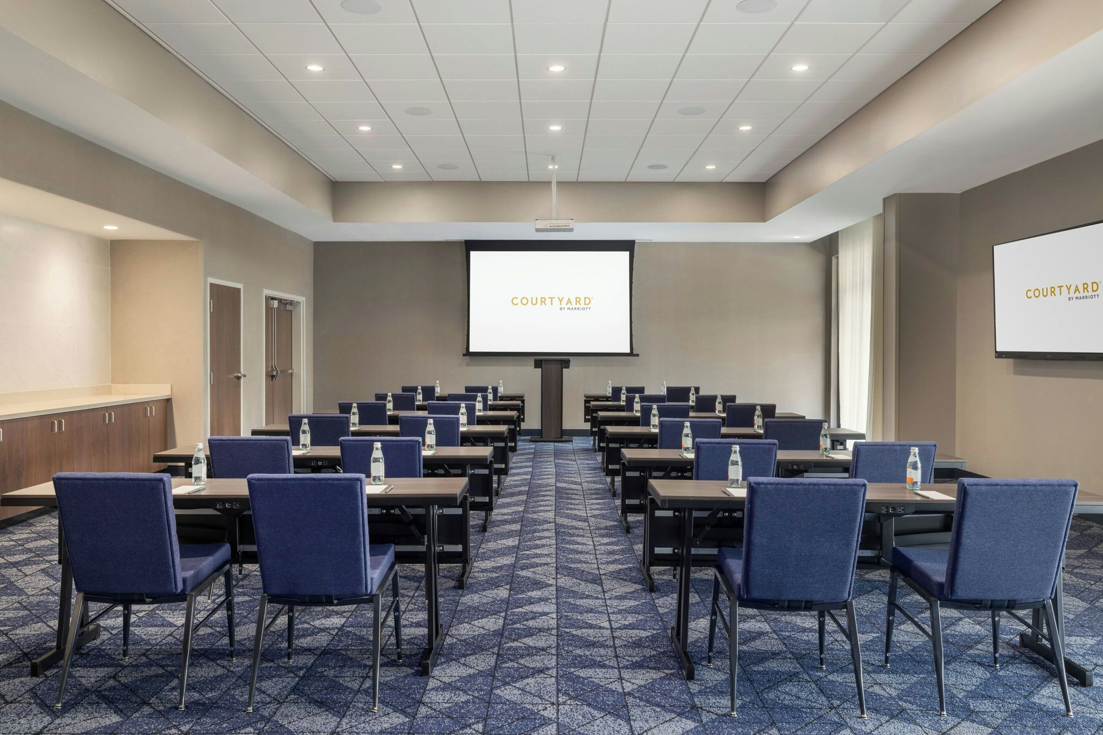 Un salón de conferencias con una pantalla grande que muestra 'COURTYARD' y una presentación, con mesas y sillas dispuestas.