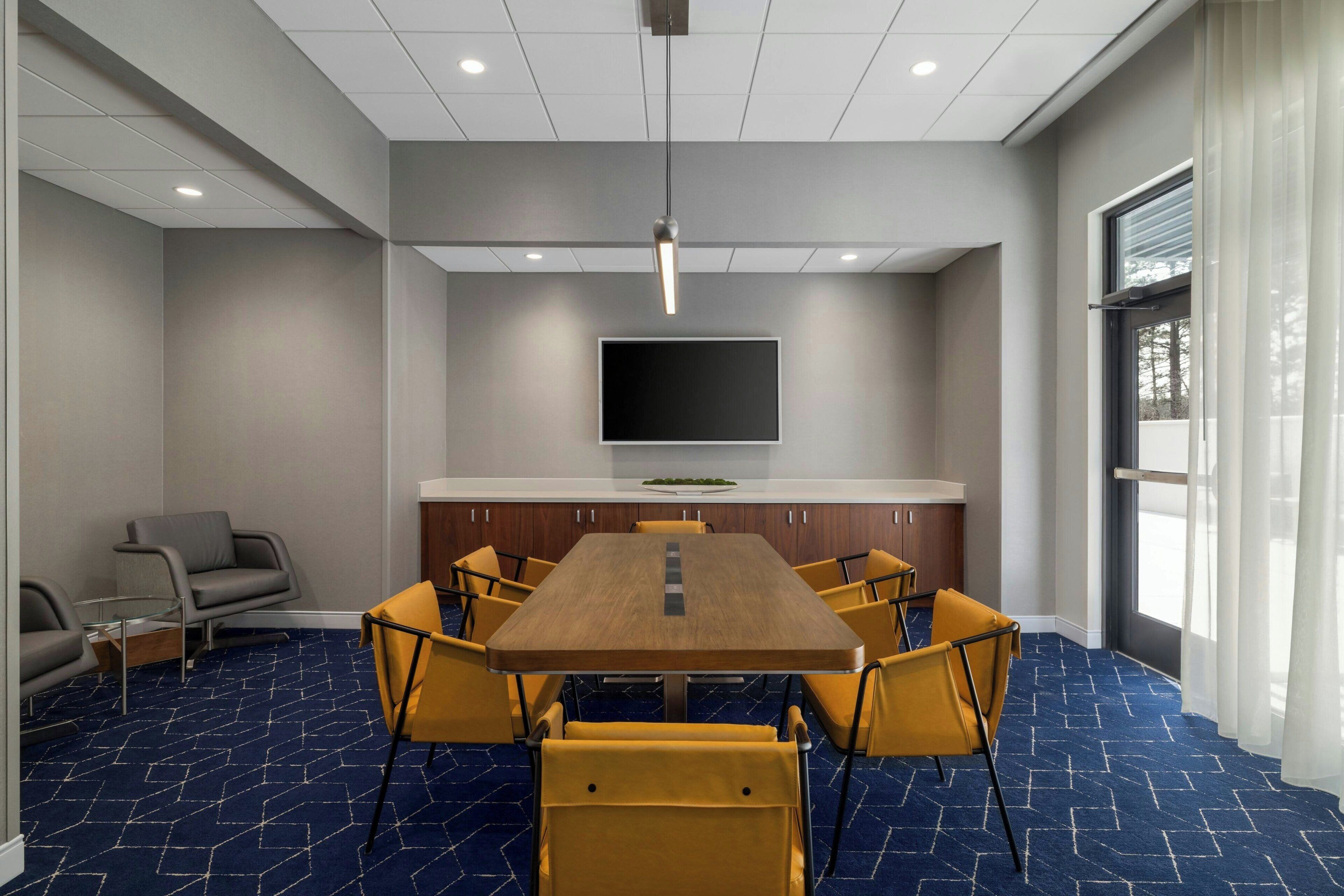 A conference room with a long table, yellow chairs, a flat-screen TV, and a window with curtains.
