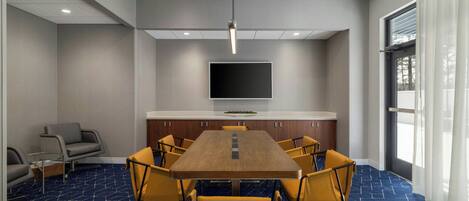 A conference room with a long table, yellow chairs, a flat-screen TV, and a window with curtains.