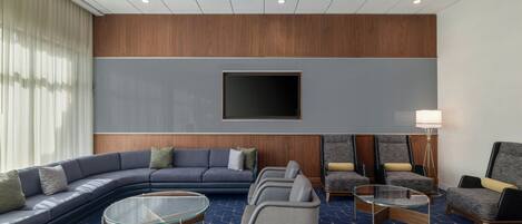 A modern hotel lobby with a curved sofa, armchairs, a glass coffee table, and a flat-screen TV mounted on the wall.