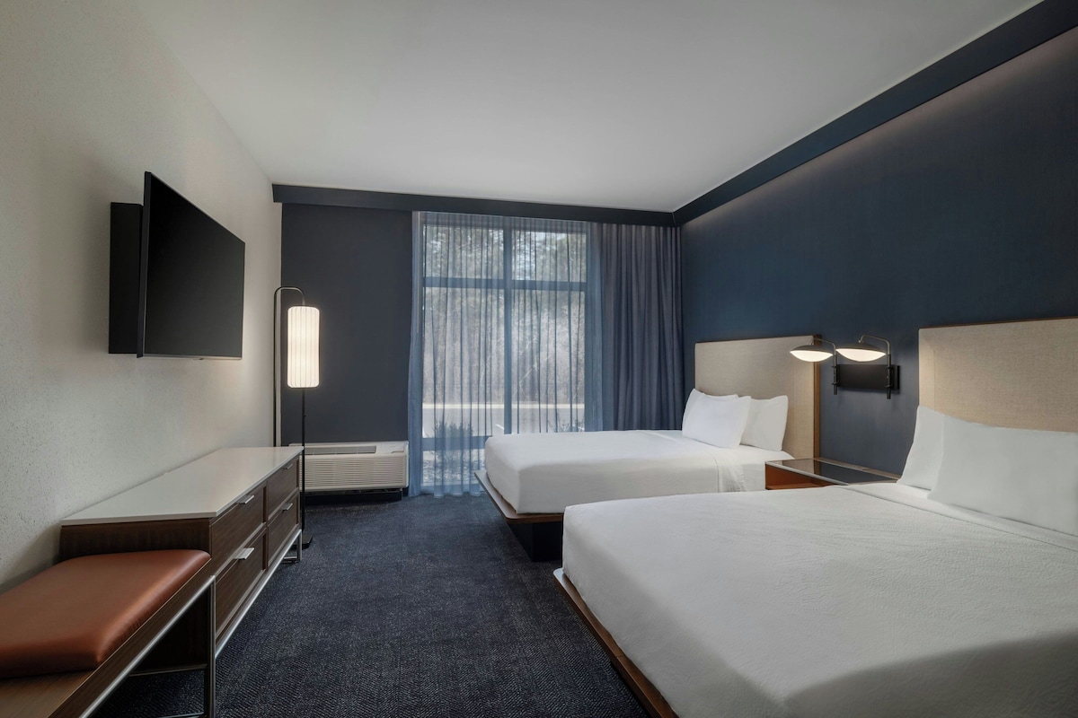 Une chambre d’hôtel avec deux lits, un bureau, une banquette, un téléviseur à écran plat et une grande fenêtre avec des rideaux.