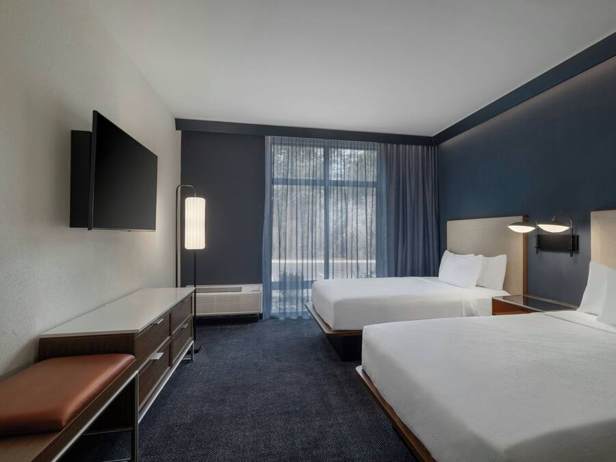 Ein Hotelzimmer mit zwei Betten, einem Schreibtisch, einer Bank, einem Flachbildfernseher und einem großen Fenster mit Vorhängen.
