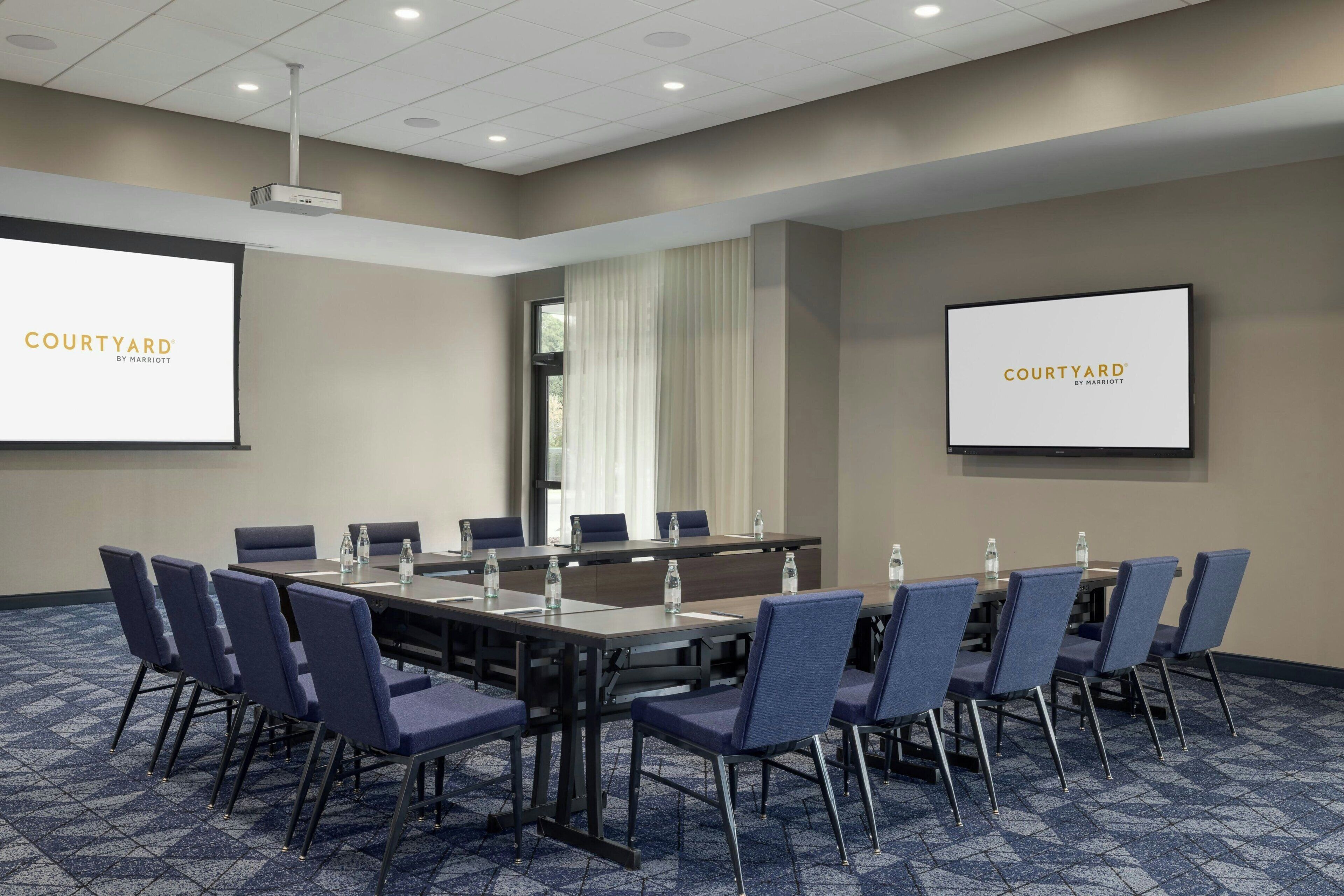 Une salle de conférence avec une longue table, des chaises bleues et deux écrans affichant le texte « COURTYARD BY MARRIOT ».