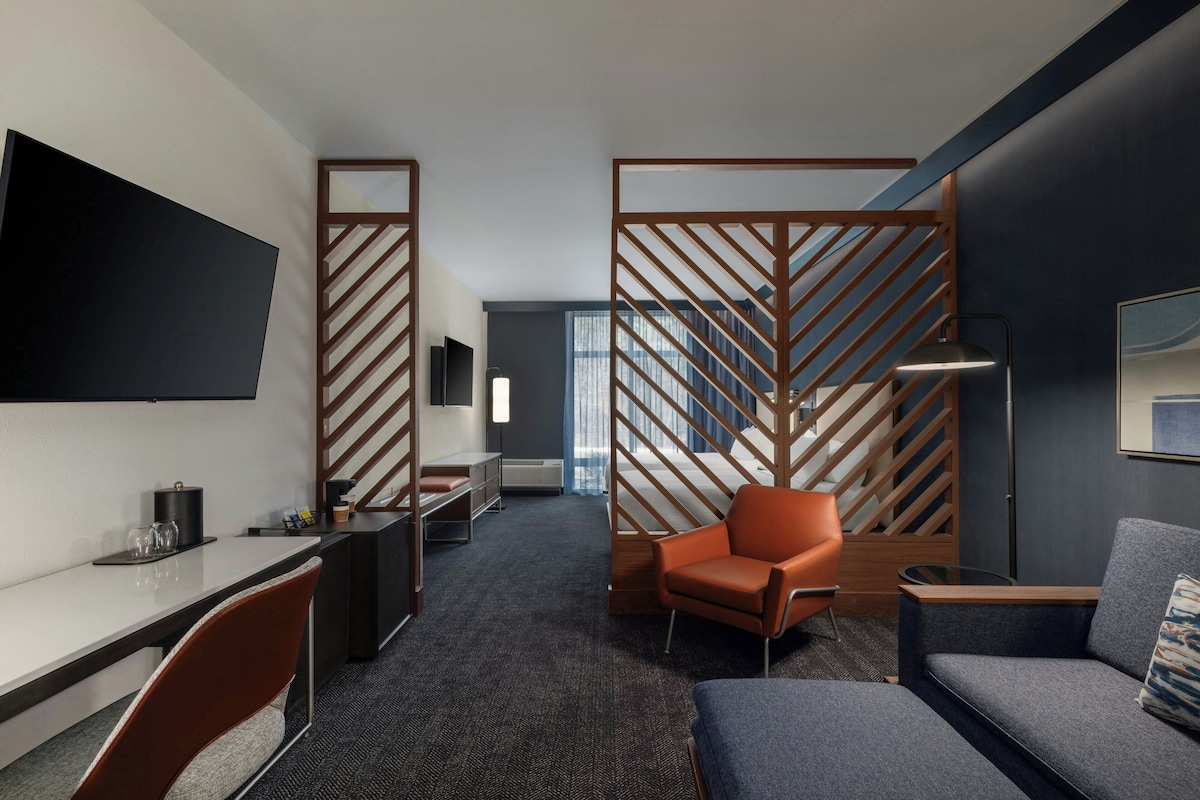 Une chambre d’hôtel équipée d’une télévision à écran plat, d’un bureau avec une chaise, d’un canapé, d’une autre chaise, d’une lampe et d’un lit séparé du reste de la pièce par une cloison en bois.