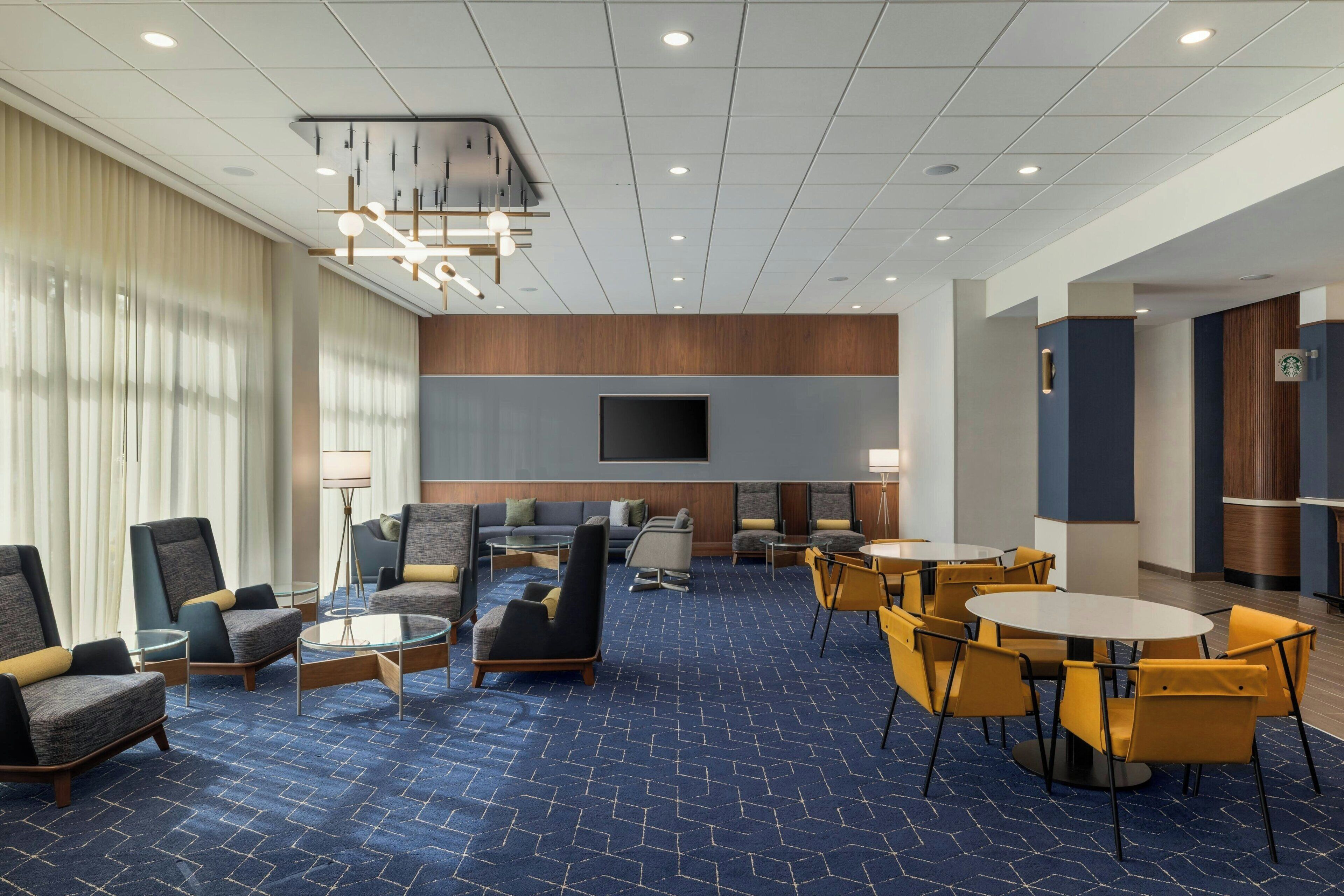 Un vestíbulo de hotel con alfombra azul, varias zonas de estar con sofás y sillones, un televisor de pantalla plana montado en la pared y un letrero de Starbucks que indica una cafetería.