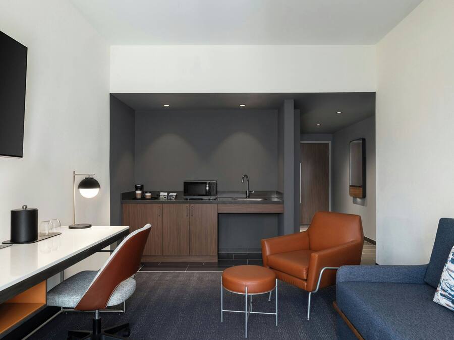 Una habitación de hotel moderna con kitchenette, un sofá, una mesa de comedor con sillas y un televisor montado en la pared.