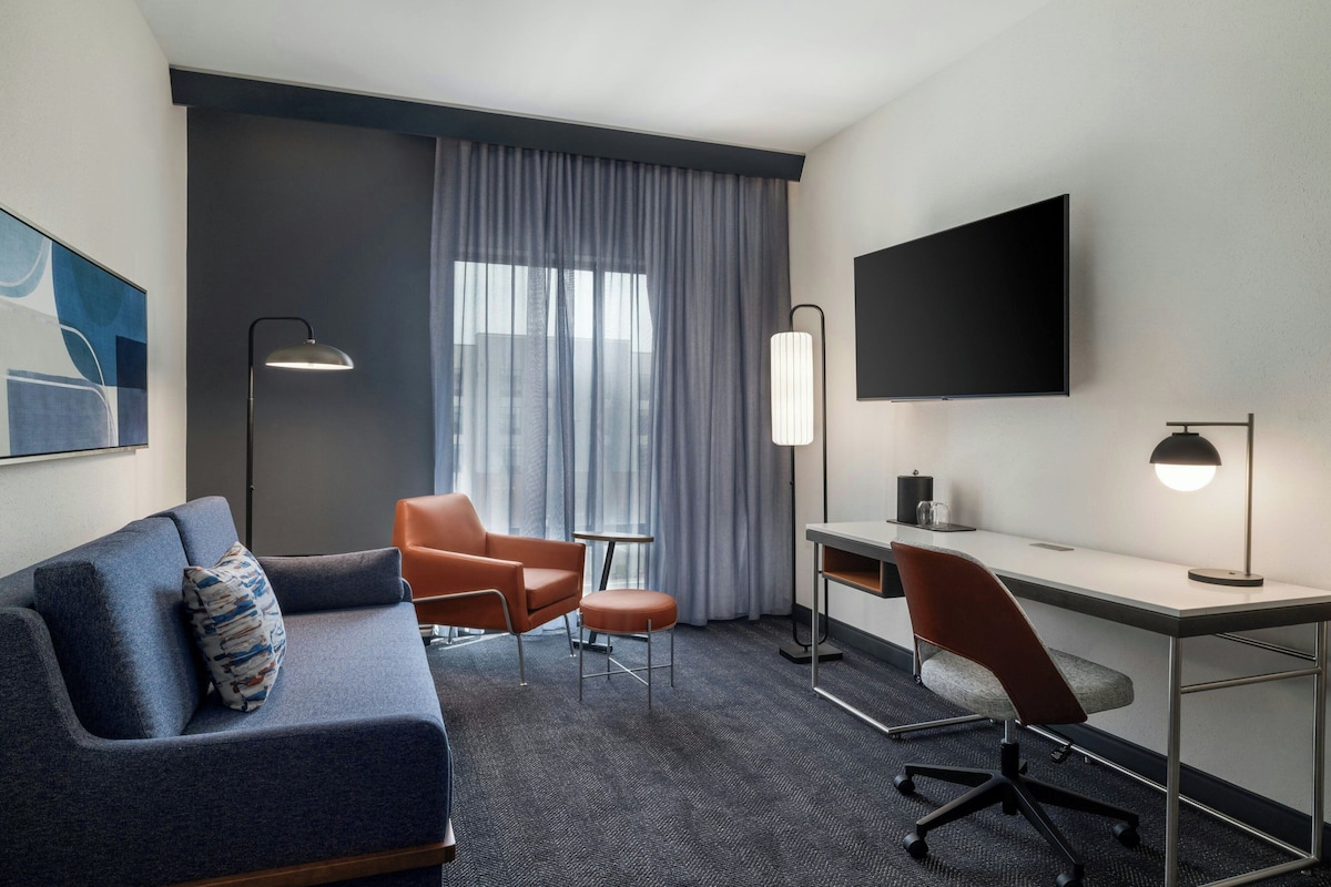 Une chambre d’hôtel avec un canapé, deux fauteuils, un bureau, une télévision et une fenêtre avec des rideaux.