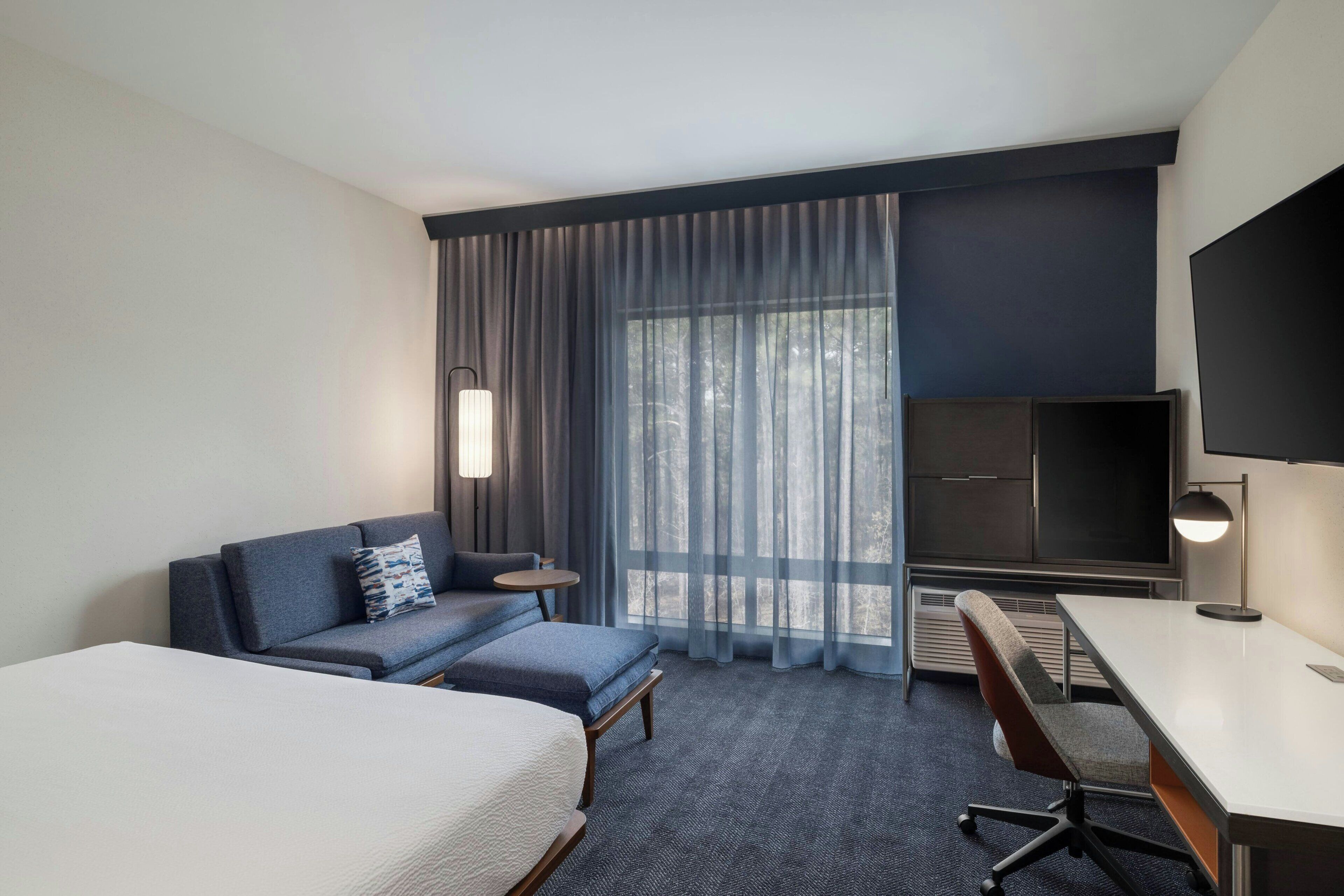A hotel room with a bed, a sofa, a desk, a chair, a TV, and a large window with curtains.