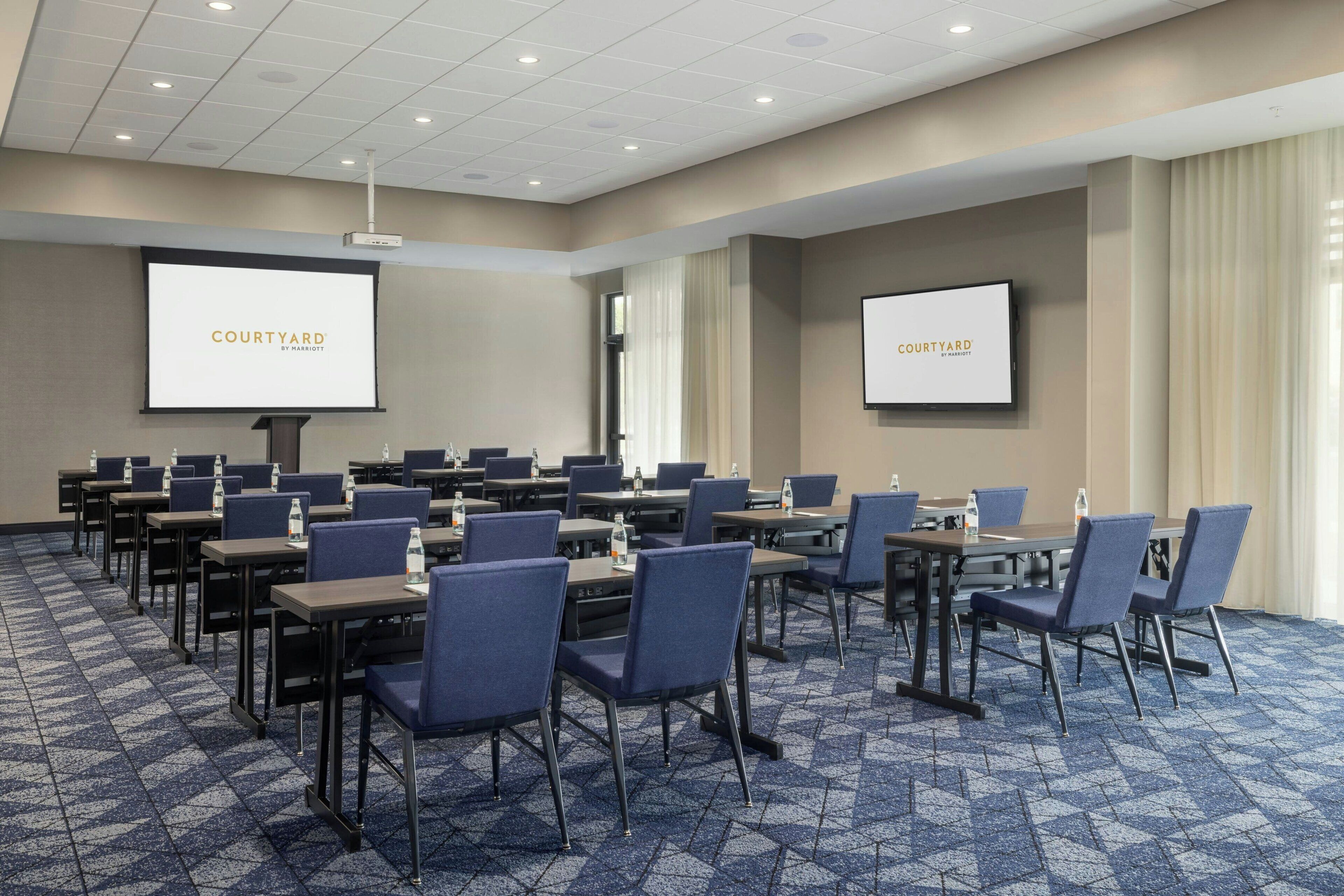 A conference room with multiple tables and chairs, a projector screen, and a large screen displaying 'COURTYARD.'