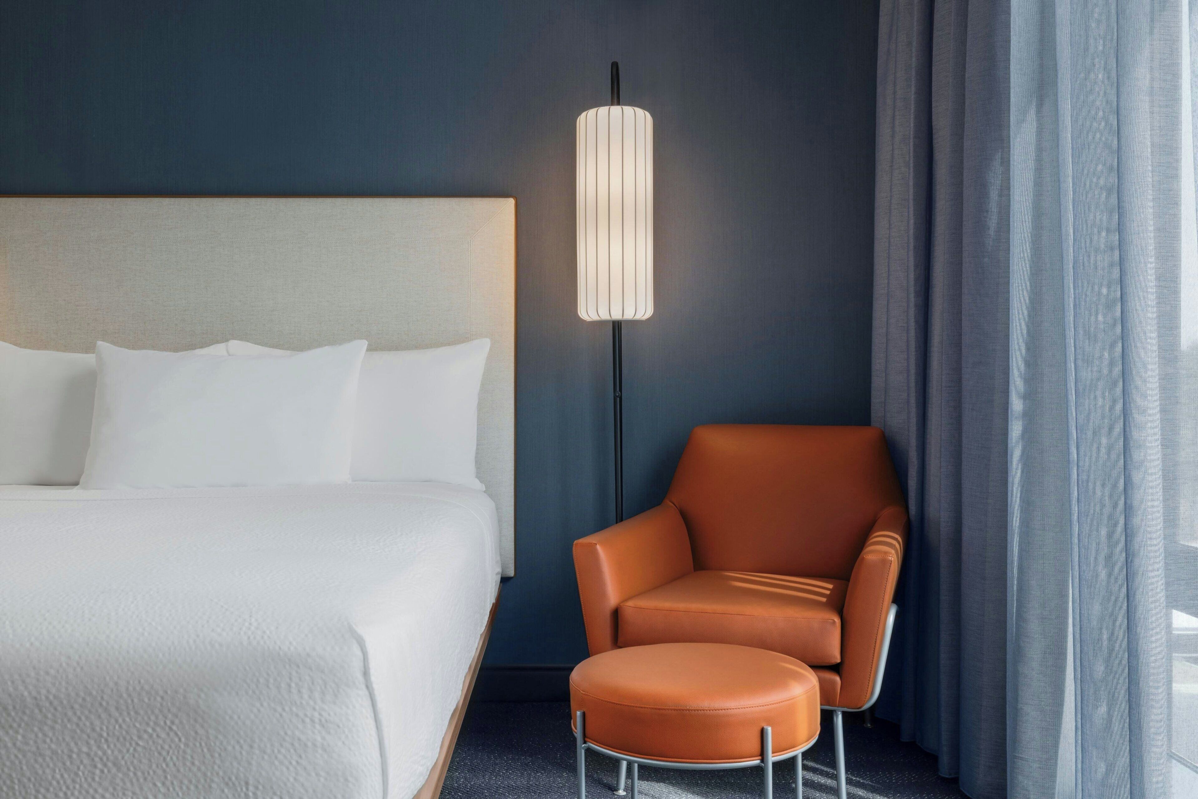 Una habitación de hotel con una cama, una silla, una lámpara de pie y un ventanal con cortinas.