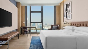 Une chambre d’hôtel avec un grand lit, un bureau avec une chaise, un banc, une télévision et une vue sur la ville.