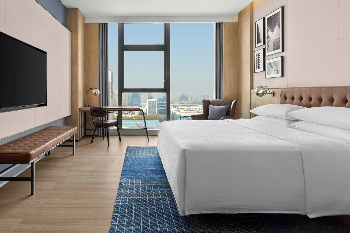 A hotel room with a large bed, a desk with a chair, a bench, a TV, and a city view.