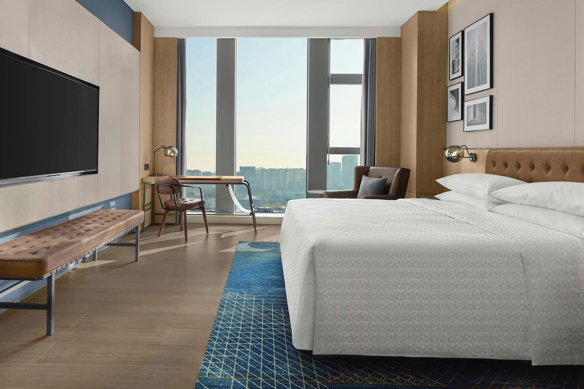 A modern hotel room with a large bed, a desk with a chair, a bench, and a large window offering a city view.
