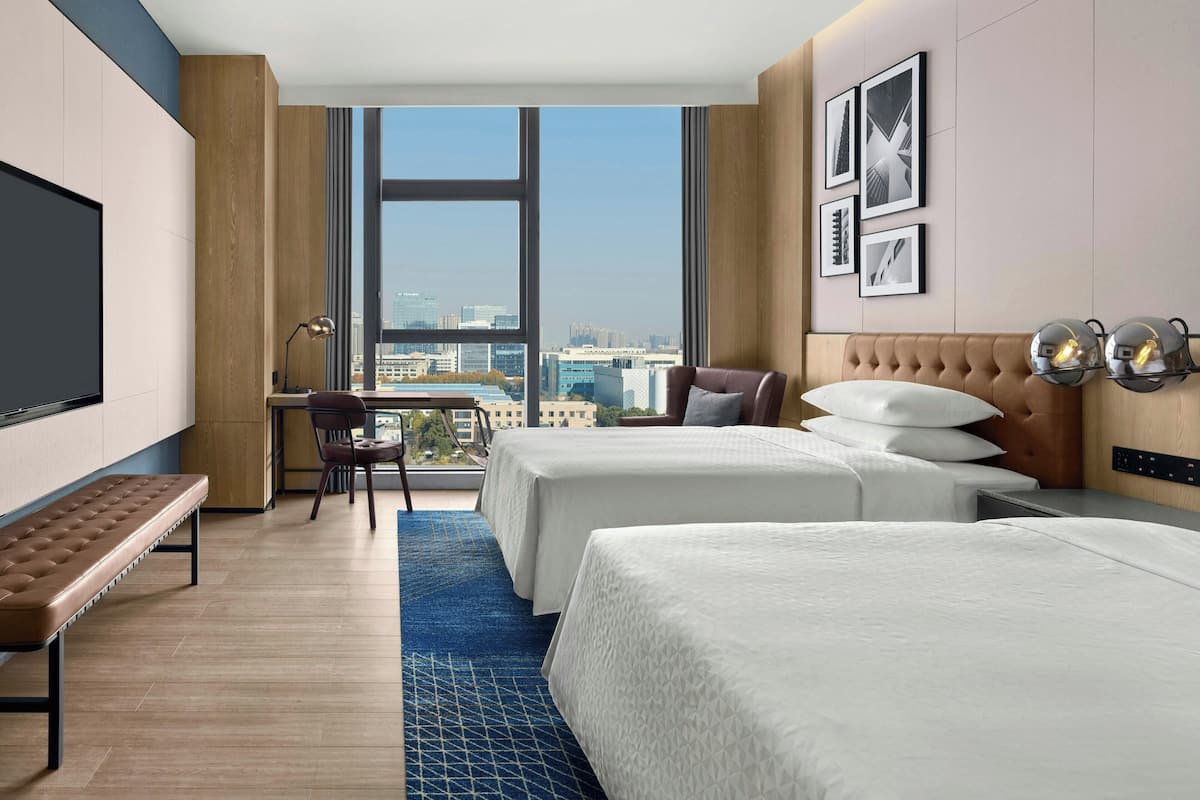 A hotel room with two beds, a desk, a chair, and a large window offering a city view.