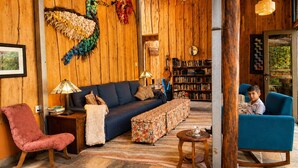 A cozy room with a wooden wall, a blue sofa, a pink chair, a bookshelf, and a child sitting on a blue chair.