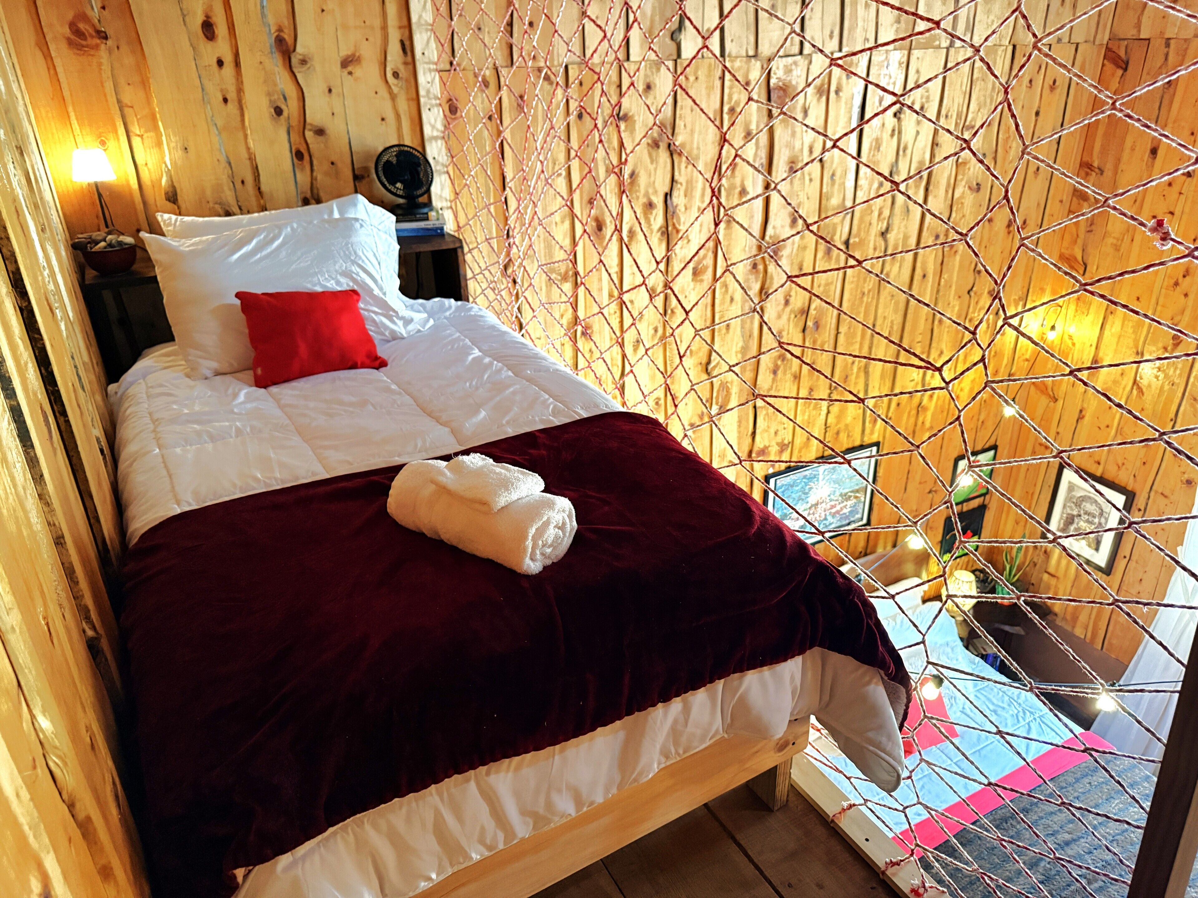 Una habitación tipo cabaña de madera con una cama, un cojín rojo, un cojín blanco, una manta roja y una toalla blanca sobre la cama.