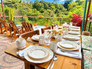 Un área de comedor al aire libre con una mesa dispuesta para una comida, rodeada de sillas y con vistas a un jardín exuberante.
