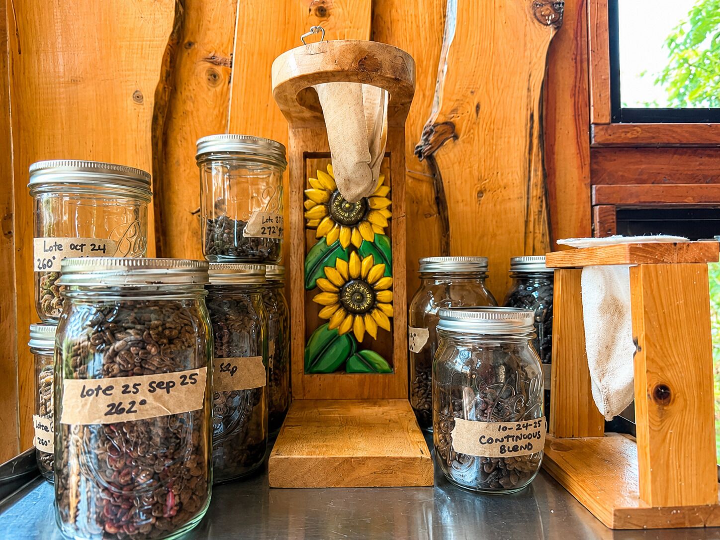 A rustic wooden interior with multiple glass jars labeled with dates and prices, a wooden spoon, and a sunflower painting.