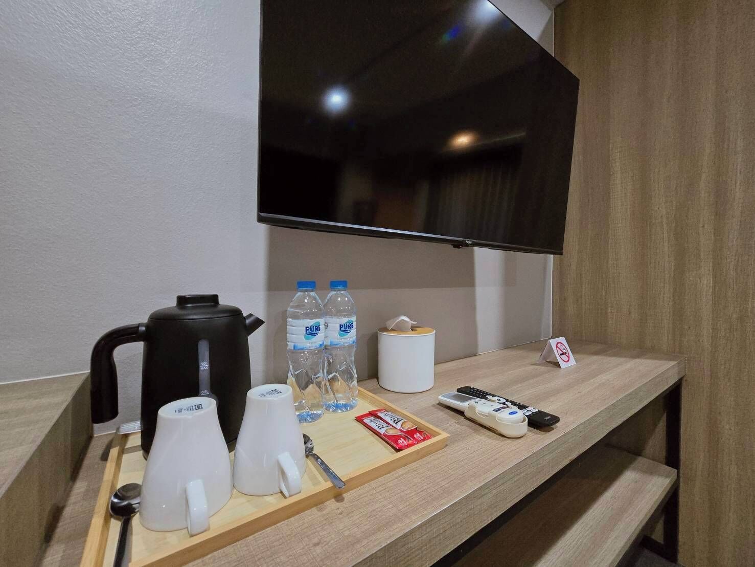 Habitación de hotel con un mostrador de madera, una cafetera negra, dos botellas de agua, un roll de pañuelos blancos, una bandeja con tazas y una cuchara, un control remoto y un televisor montado en la pared.