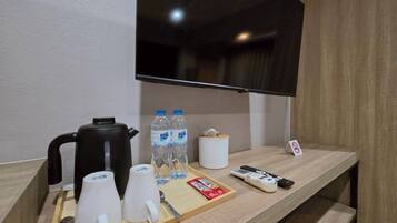 Habitación de hotel con un mostrador de madera, una cafetera negra, dos botellas de agua, un roll de pañuelos blancos, una bandeja con tazas y una cuchara, un control remoto y un televisor montado en la pared.
