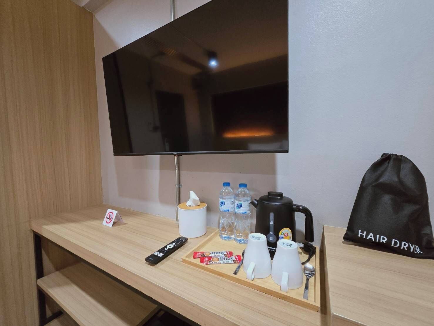 A hotel room with a TV mounted on the wall, a tray table with a remote, water bottles, a kettle, and a hair dryer bag.