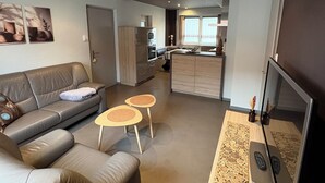Una sala moderna con un sofá, dos mesitas y un televisor grande. Se observa una cocina al fondo con armarios y una ventana.